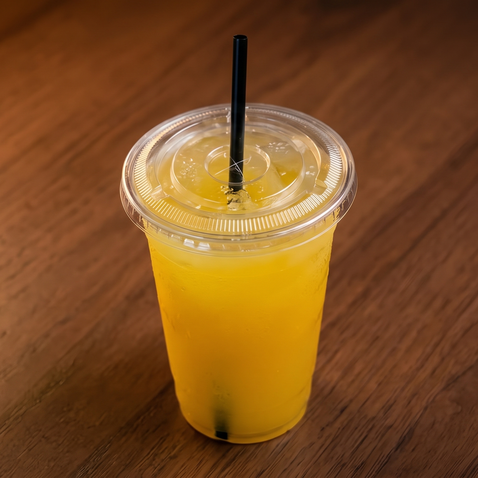 Bright yellow iced drink in a clear plastic cup with a black straw on a wooden table.